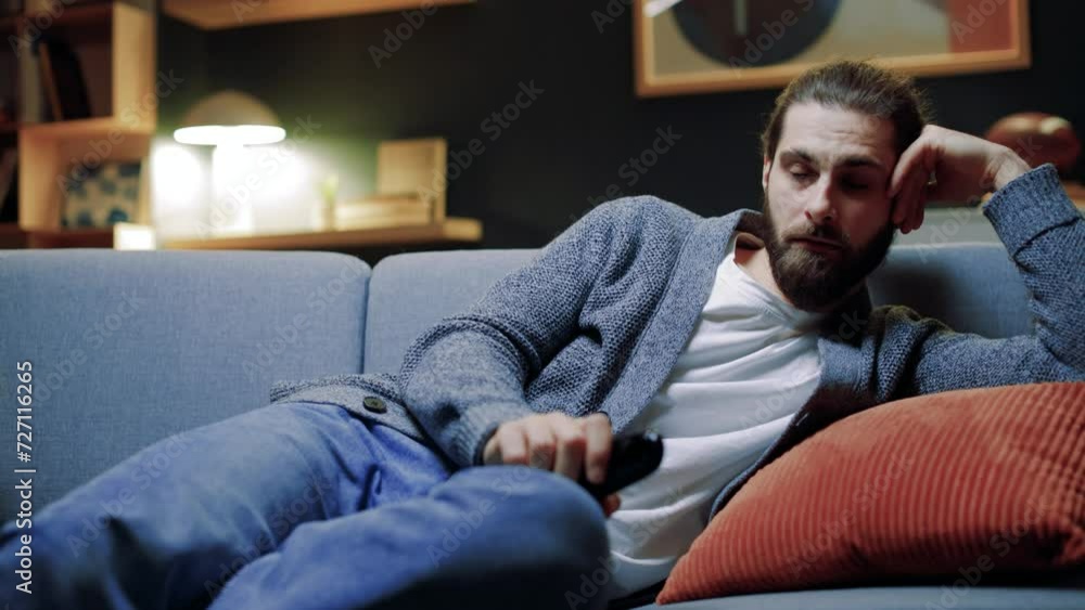 Portrait of tired young bearded man with remote controller switches channels on TV trying to choose something interesting looking at screen at late night. Boring TV shows films movie Portrait of tired young bearded man with remote controller switches channels on TV trying to choose something interesting looking at screen at late night. Boring TV shows films movie