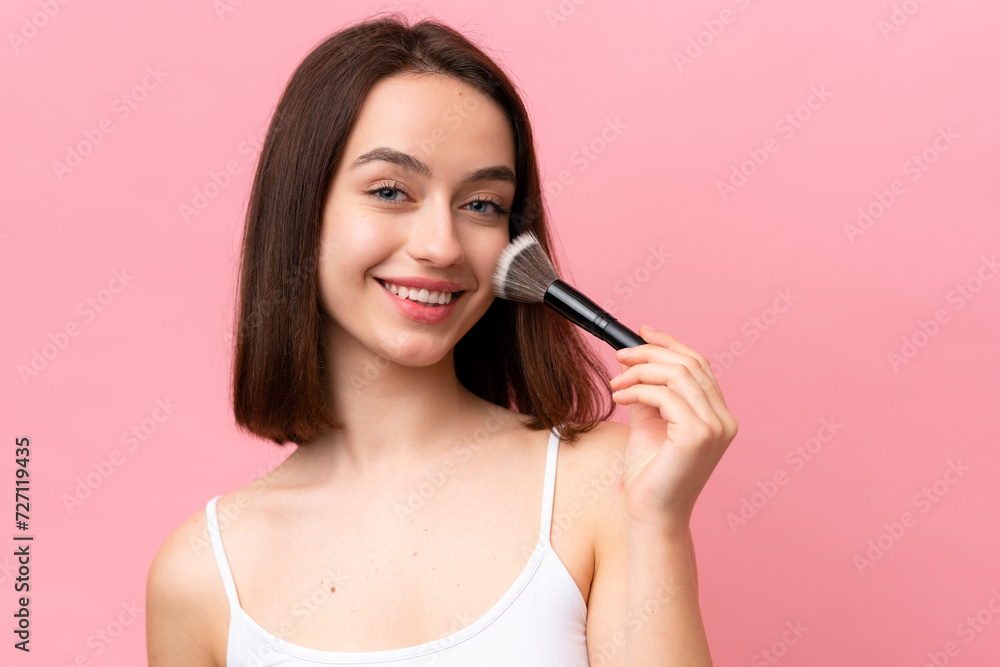 Young Ukrainian woman isolated on pink background holding makeup brush and whit happy expression