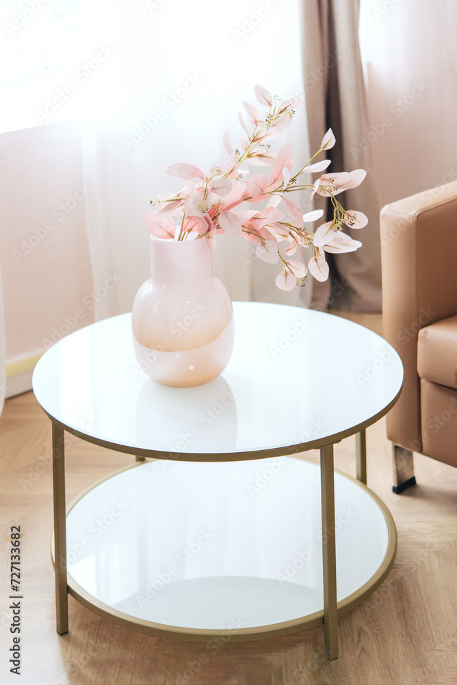 Branches of pink flowers in glass vase on small white glass table against window, interior details with armchair and curtains