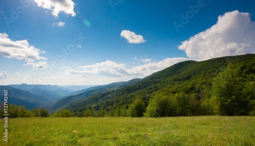 mountainous rural landscape on a sunny afternoon forested hills and green grassy meadows in evening light ridge in the distance sunny weather with fluffy clouds on the bright blue sky © Emanuel