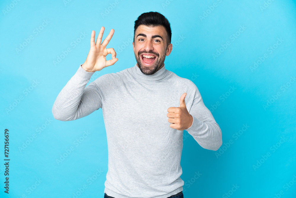 Caucasian man over isolated blue background showing ok sign and thumb up gesture Caucasian man over isolated blue background showing ok sign and thumb up gesture