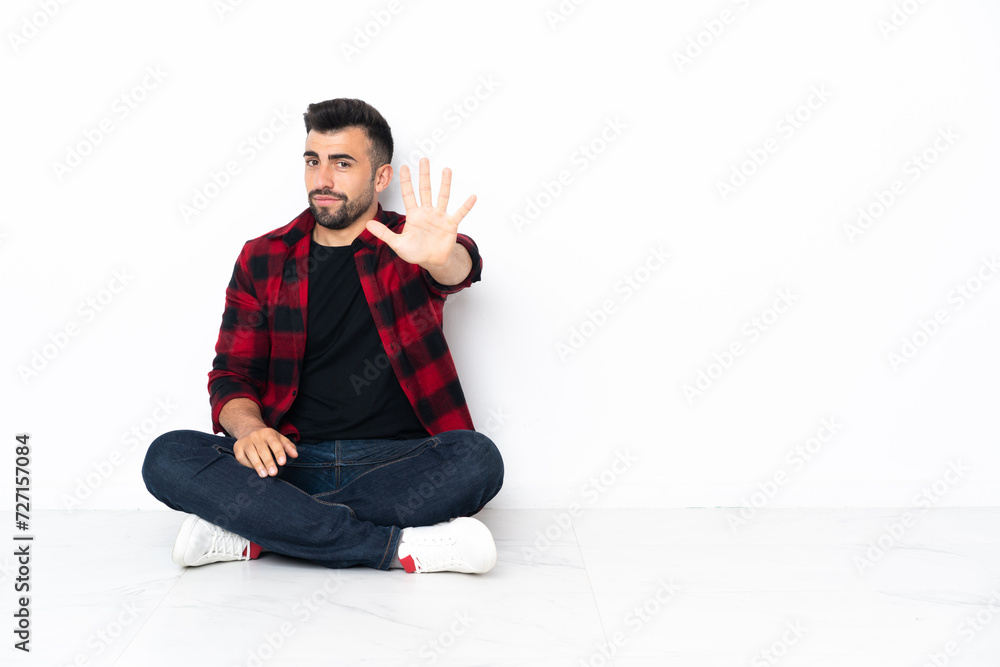 Young handsome man sitting on the floor counting five with fingers Young handsome man sitting on the floor counting five with fingers