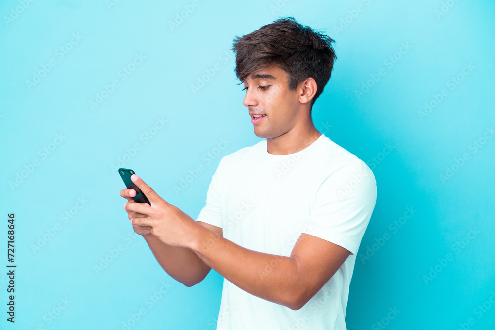 Young caucasian man isolated on blue background sending a message or email with the mobile Young caucasian man isolated on blue background sending a message or email with the mobile