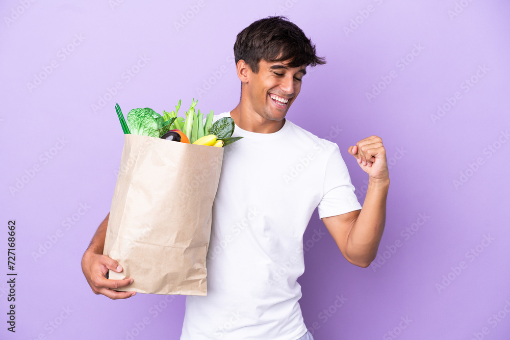 Young man holding a grocery shopping bag isolated on purple background celebrating a victory Young man holding a grocery shopping bag isolated on purple background celebrating a victory