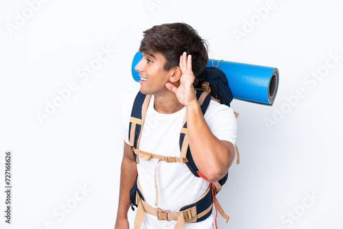 Young mountaineer woman with a big backpack over isolated white background listening to something by putting hand on the ear