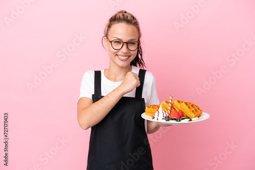 Restaurant waiter Russian girl holding waffles isolated on pink background celebrating a victory