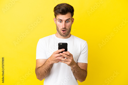 Young caucasian handsome man isolated on yellow background looking at the camera while using the mobile with surprised expression © luismolinero