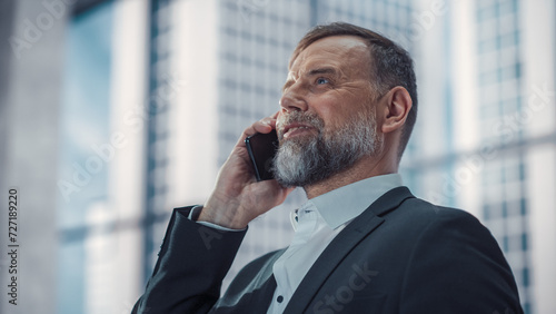 Confident Middle Aged Businessman in a Suit Standing in Modern Office, Answering a Phone Call, Looking Out of the Window on Big City with Skyscrapers. Successful Company CEO Planning Work Projects