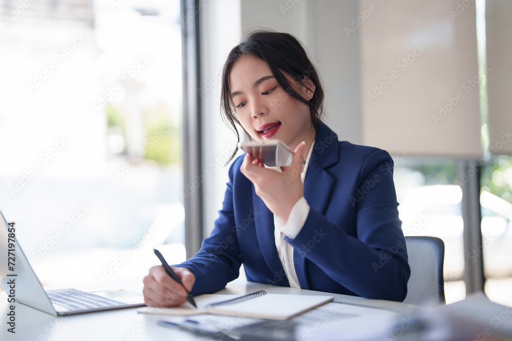 Businesswomen using smartphone to call with customer about financial and investment data of new business and taking notes while working to analysis strategy about new startup project in workplace Businesswomen using smartphone to call with customer about financial and investment data of new business and taking notes while working to analysis strategy about new startup project in workplace