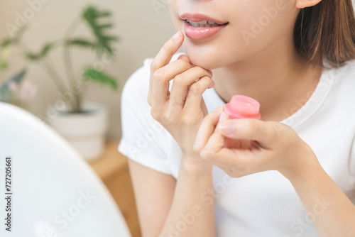 Lip care concept, happy asian young woman finger touching lips in front of mirror at home after applying lipstick balm on dry mouth from natural beauty product, skincare routine, makeup and cosmetics. photo