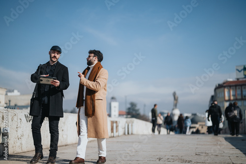 Two stylish men discussing business on a city bridge on a sunny day.