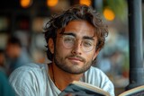 A handsome young man in stylish eyewear, holding a book, exudes confidence in a cafe.