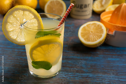 Glass of lemonade with slices and mint inside, on a rustic table with lemons, sugar and a manual juicer, close-up.