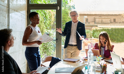 Mature businessman with document gesticulating and speaking to multiracial colleagues while standing against window in modern workplace photo