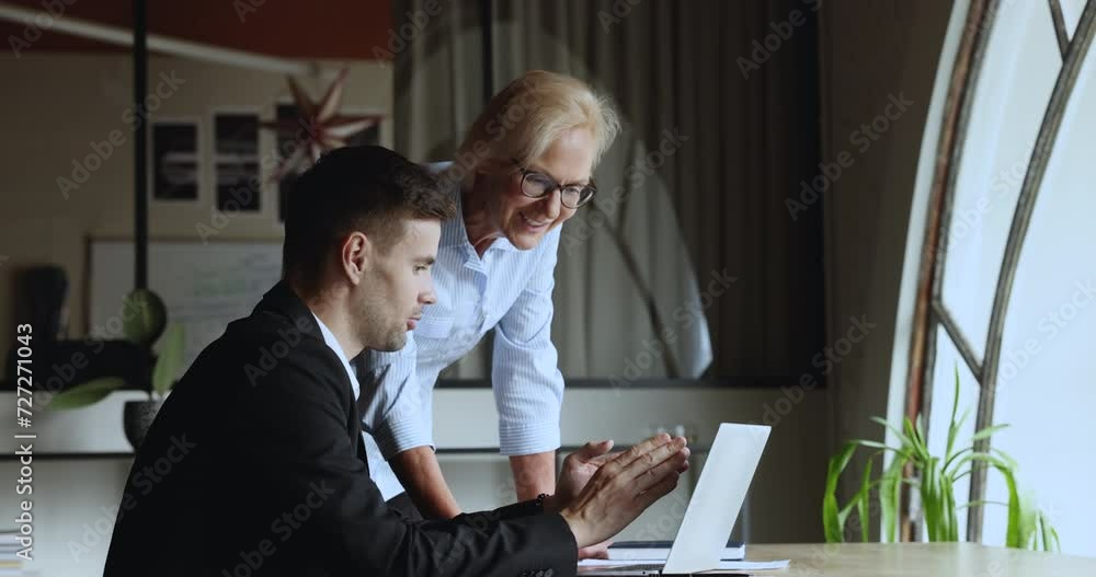 Positive motivated young manager man showing online project presentation to happy senior female boss, sitting at laptop, pointing at screen, speaking to mature businesswoman Positive motivated young manager man showing online project presentation to happy senior female boss, sitting at laptop, pointing at screen, speaking to mature businesswoman