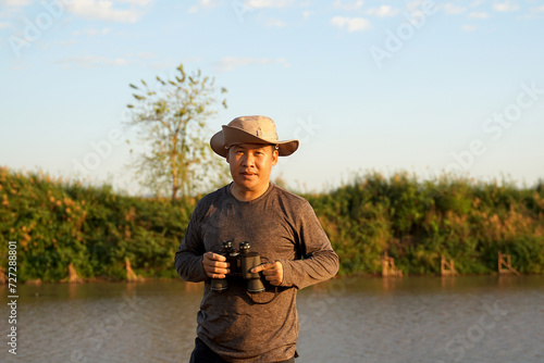 Asian man uses binoculars to observe local birds along a river. Soft and selective focus. © Aoy_Charin