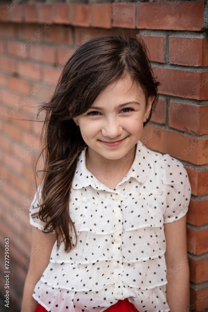 Front portrait of a cheerful cute little girl in blouse posing near brick wall, looking at camera. Vertical v Front portrait of a cheerful cute little girl in blouse posing near brick wall, looking at camera. Vertical v