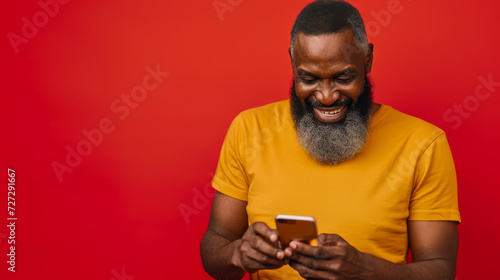 bearded older man with a top knot hairstyle, laughing and looking at his phone, wearing a yellow t-shirt against a solid red background © MP Studio