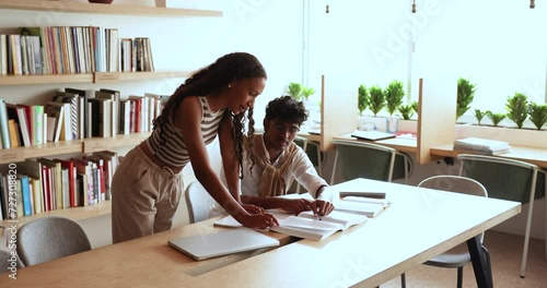 Young African female teacher or schoolmate provide explanation to Indian teenager student guy, explain task to-do at meeting in library. Education, apprenticeship, studying process, additional classes photo