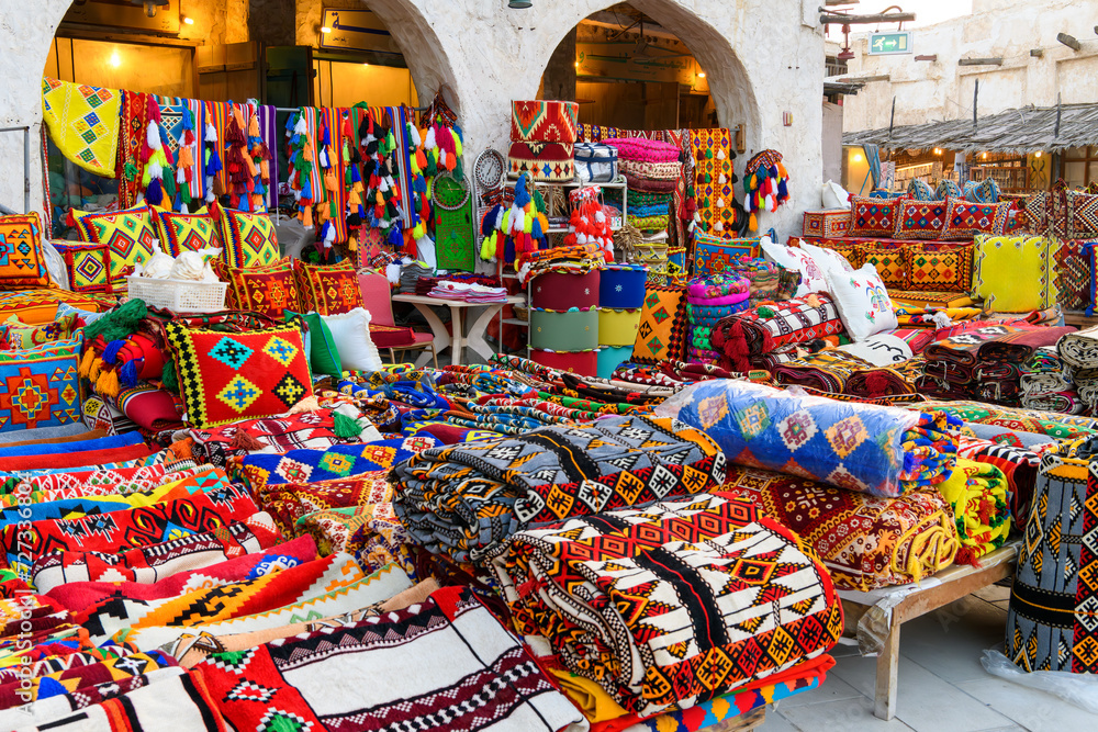 A colorful outdoor storefront display selling vibrantly colored fabrics on blankets, rugs and pillows in the Souq Waqif marketplace of Doha, Qatar.