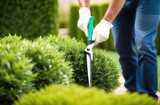 A gardener in blue work pants and white gloves meticulously trims a green bush with manual hedge clippers in a vibrant garden setting