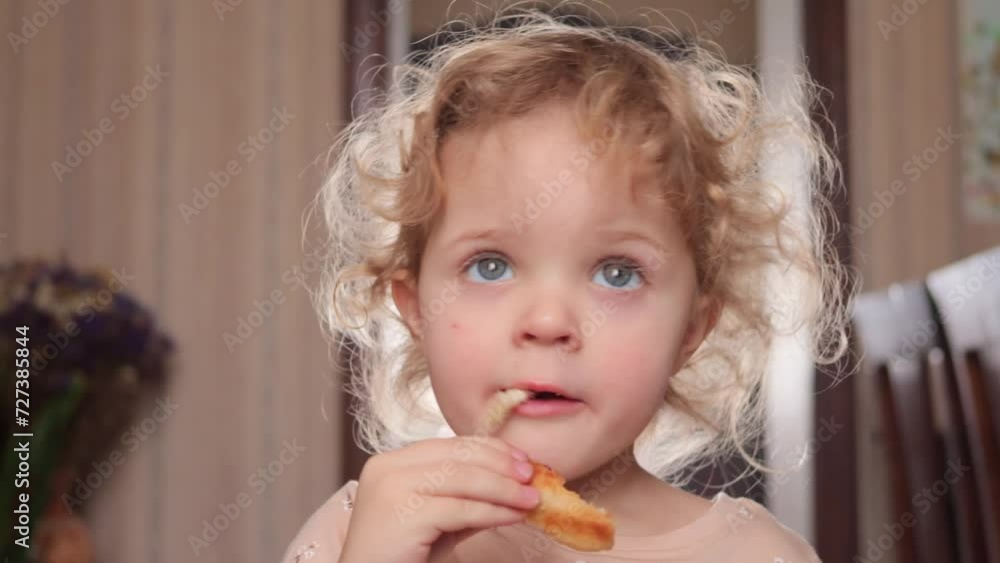 Beautiful blonde wavy haired toddler child girl eating cookies or pie while standing in home interior Beautiful blonde wavy haired toddler child girl eating cookies or pie while standing in home interior