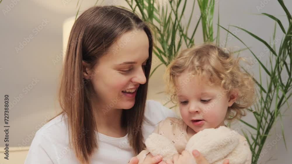Attractive beautiful woman with her infant daughter enjoying playful time together at home talking while playing with fluffy rabbit on weekend Attractive beautiful woman with her infant daughter enjoying playful time together at home talking while playing with fluffy rabbit on weekend