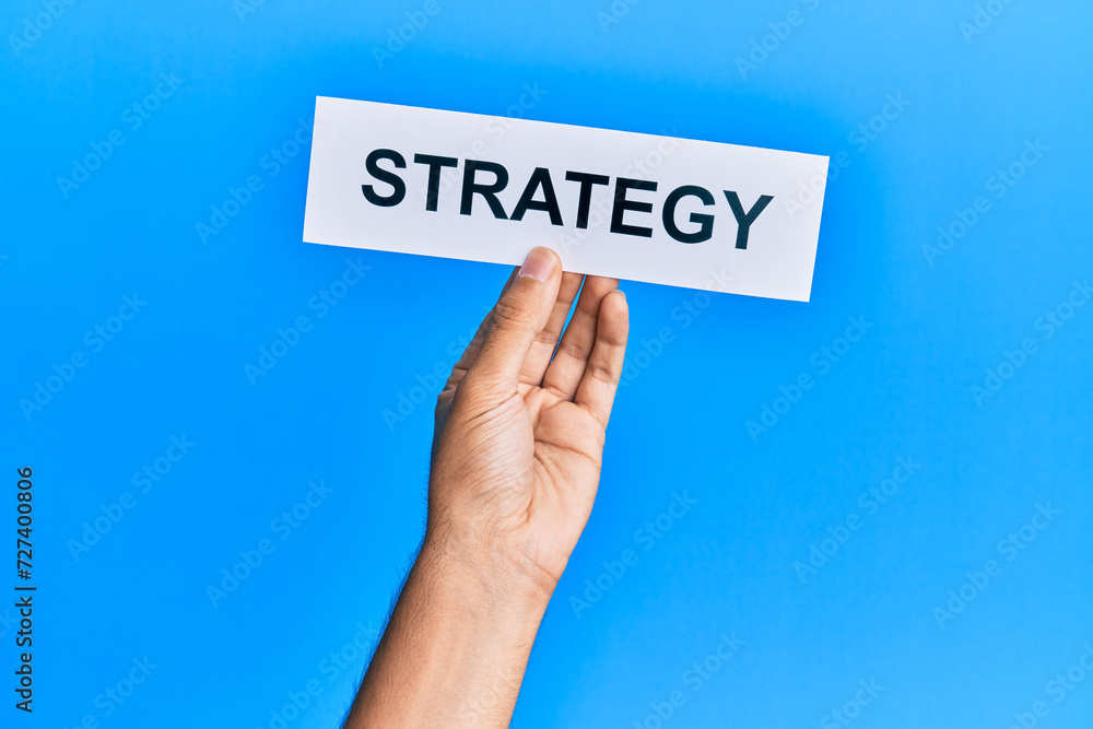 Hand of caucasian man holding paper with strategy word over isolated blue background Hand of caucasian man holding paper with strategy word over isolated blue background