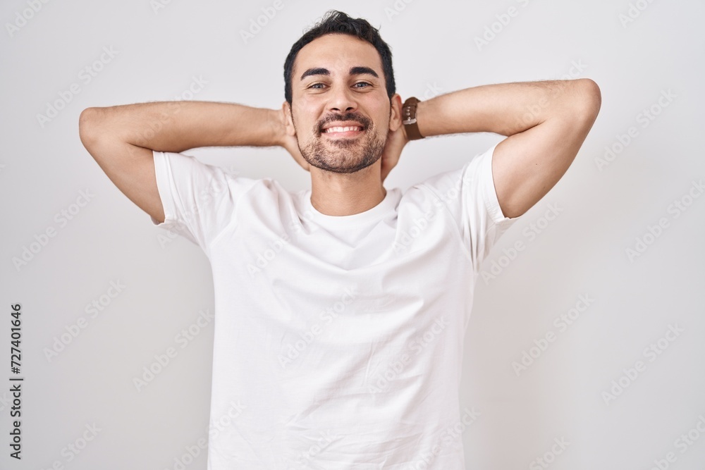 Handsome hispanic man standing over white background relaxing and stretching, arms and hands behind head and neck smiling happy Handsome hispanic man standing over white background relaxing and stretching, arms and hands behind head and neck smiling happy