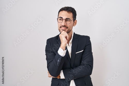 Handsome business hispanic man standing over white background with hand on chin thinking about question, pensive expression. smiling and thoughtful face. doubt concept.