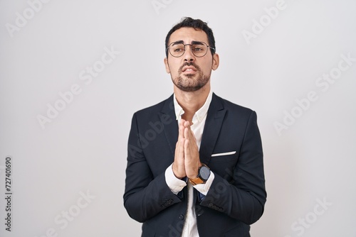 Handsome business hispanic man standing over white background begging and praying with hands together with hope expression on face very emotional and worried. begging.