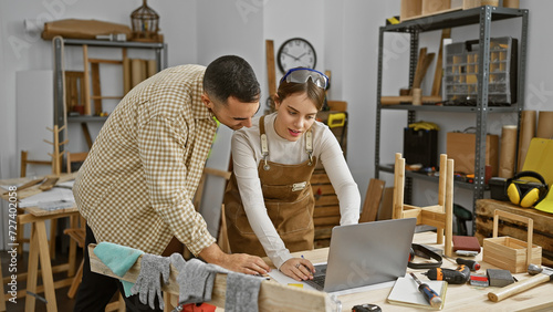 A man and woman collaborate in a carpentry workshop, reviewing designs on a laptop surrounded by woodworking tools.