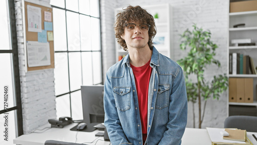 A handsome young man with curly hair and glasses posing confidently in a modern office setting.