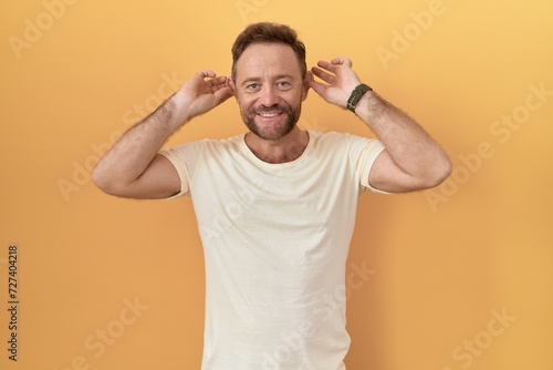 Middle age man with beard standing over yellow background smiling pulling ears with fingers, funny gesture. audition problem