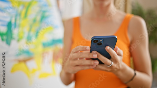 A young woman in an orange top uses a smartphone in a creative studio space with artwork in the background. photo