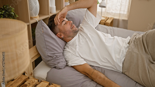 Attractive young hispanic man, stressed and exhausted, lying in bed, resting but awakened by troubling headache in his cozy bedroom, a picture of worry and anxiety