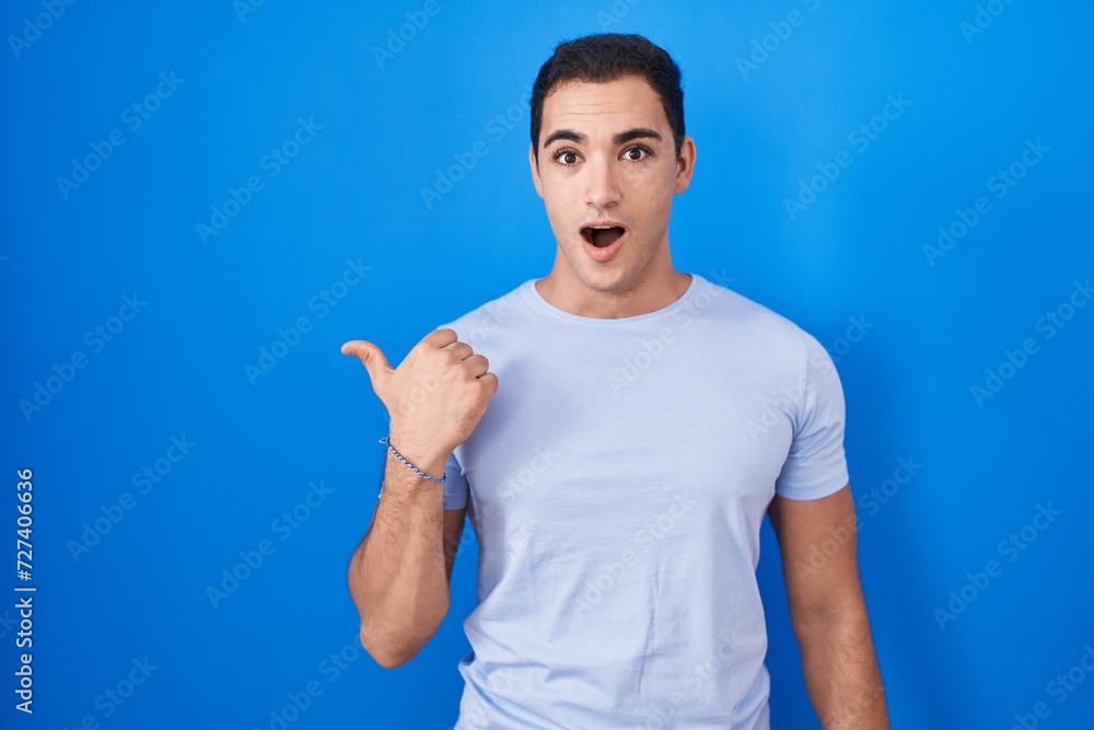 Young hispanic man standing over blue background surprised pointing with hand finger to the side, open mouth amazed expression. Young hispanic man standing over blue background surprised pointing with hand finger to the side, open mouth amazed expression.