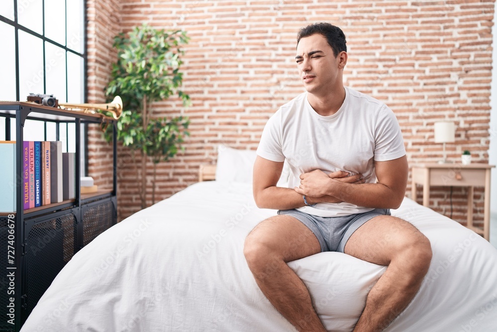 Young hispanic man suffering for stomach ache sitting on bed at bedroom Young hispanic man suffering for stomach ache sitting on bed at bedroom