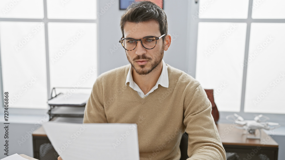 Fototapeta premium Handsome hispanic man with beard reading documents in modern office setting