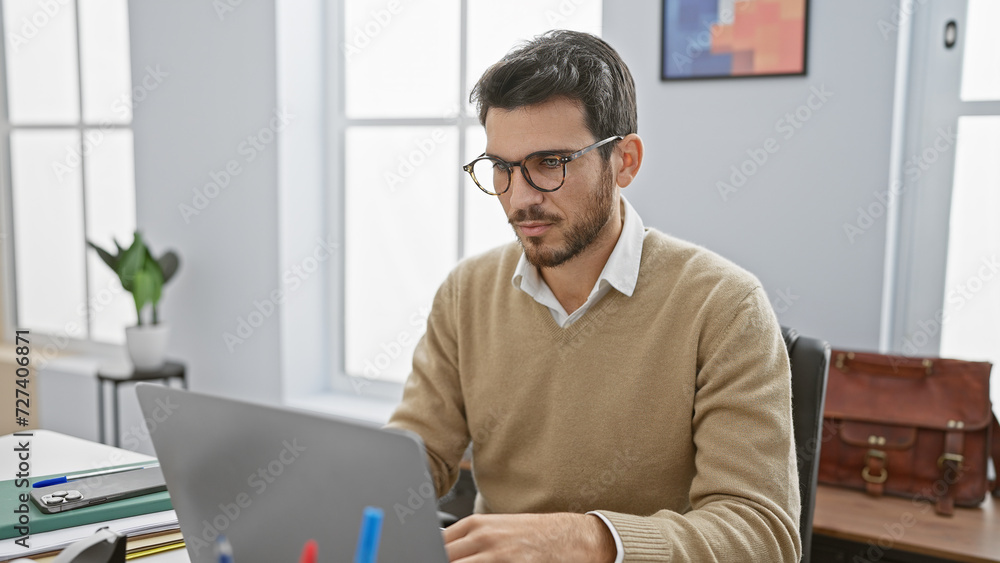 Handsome hispanic man with beard wearing glasses working on laptop in modern office setting