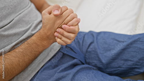 Closeup of a young man's clasped hands resting on a bed in an indoor bedroom setting reflecting anticipation or concern.