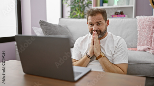A hopeful middle-aged hispanic man with grey hair in a home setting using a laptop on a sofa.