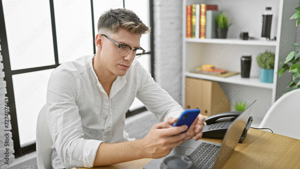 Handsome young caucasian man, a serious business worker, engrossed in his thoughts at office, working hard while typing a message on laptop, doubting a business idea, looking at smartphone screen Handsome young caucasian man, a serious business worker, engrossed in his thoughts at office, working hard while typing a message on laptop, doubting a business idea, looking at smartphone screen