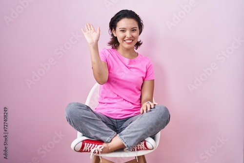 Hispanic young woman sitting on chair over pink background waiving saying hello happy and smiling, friendly welcome gesture