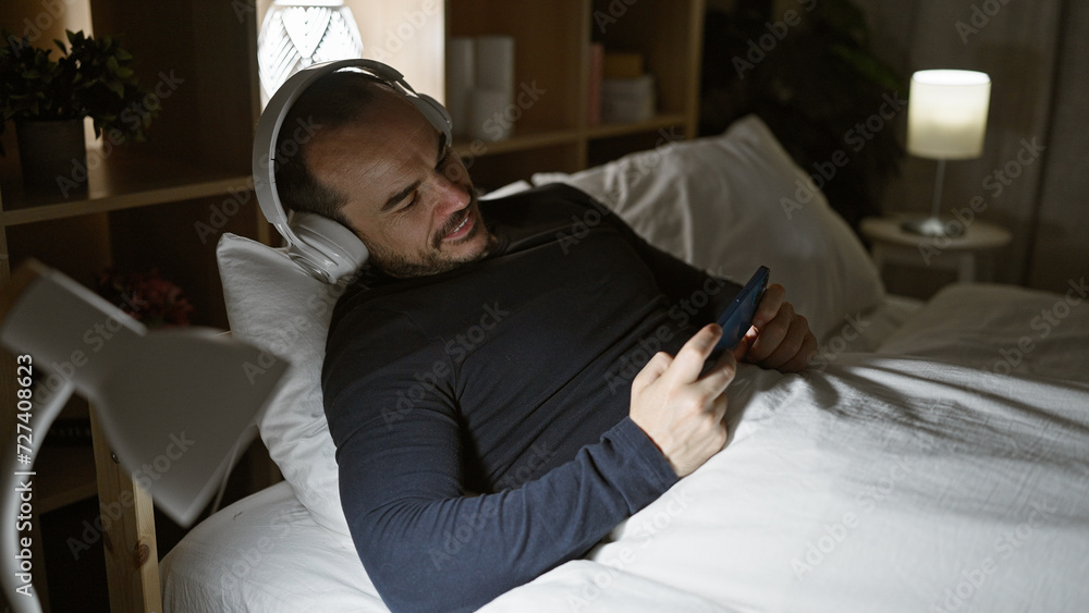 Hispanic man with beard enjoying music on headphones while holding smartphone in bedroom at night. Hispanic man with beard enjoying music on headphones while holding smartphone in bedroom at night.