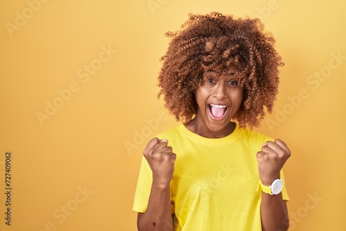Young hispanic woman with curly hair standing over yellow background celebrating surprised and amazed for success with arms raised and open eyes. winner concept.