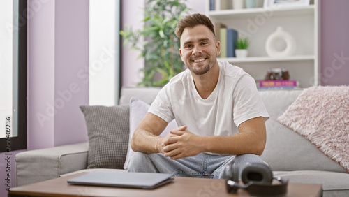 Handsome young hispanic man smiling in a modern living room with a couch and decorations