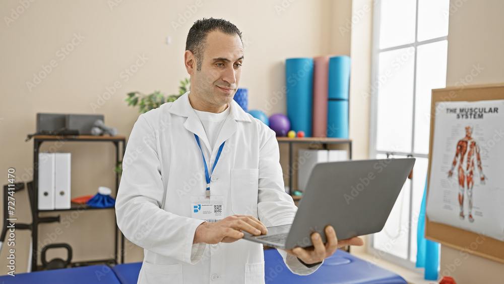 A hispanic man works on a laptop in a bright physical therapy clinic, exemplifying professionalism and healthcare.