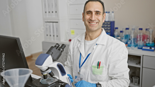 Smiling hispanic man in lab with microscope, clinic scientist in white coat.