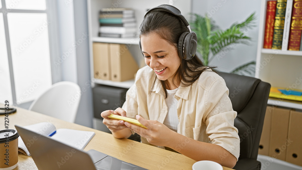 Smiling young hispanic woman worker enjoying video on her smartphone, using headphones in a bustling office, exhibiting a beautiful mix of elegance and professional success. Smiling young hispanic woman worker enjoying video on her smartphone, using headphones in a bustling office, exhibiting a beautiful mix of elegance and professional success.